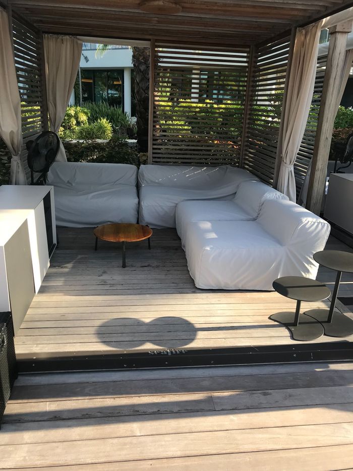 A shaded outdoor seating area with white couches, wooden floors, and a small round table.
