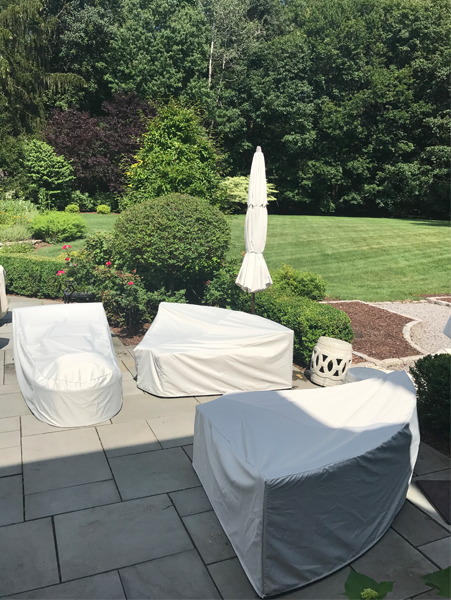 Patio furniture covered in white, with a closed umbrella, on a stone patio, with a green lawn and trees in the background.