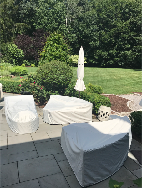 Outdoor patio furniture covered with beige protective covers on a sunny day; garden and trees in the background.