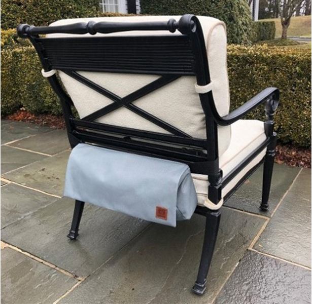 A black wrought-iron chair with cream cushions has a gray bag attached to the back. The chair sits on a stone patio.
