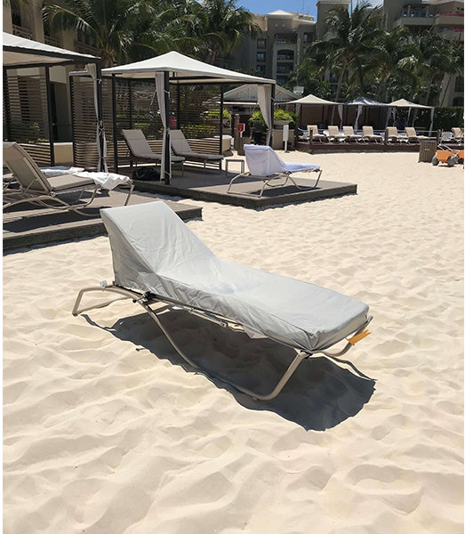 Beach chair covered in grey fabric on a sandy beach. Other chairs, cabanas, and resort buildings are in the background.