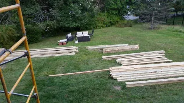 Lumber laid out on green grass, ready for construction. A yellow ladder is visible in the foreground.