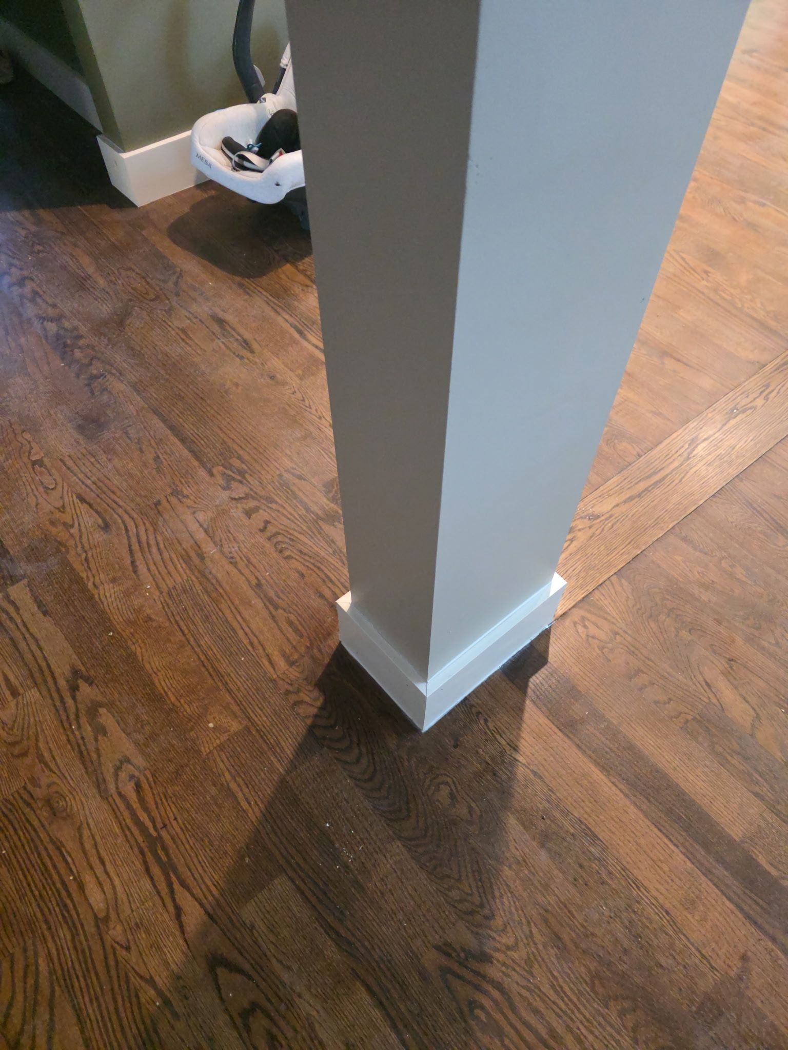 A square white column with baseboard on dark hardwood floor, casting a shadow.