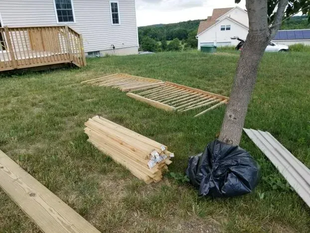 Lumber, lattice, and trash bag near a tree in a grassy yard, with a house and deck in the background.