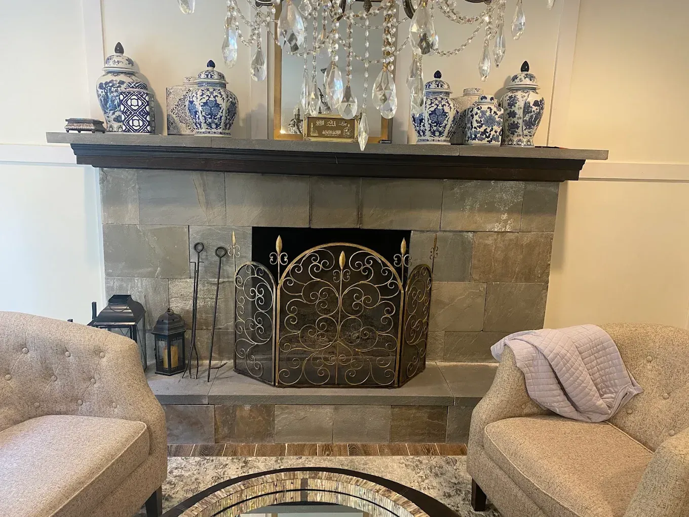 Fireplace with blue and white vases, flanked by upholstered chairs and a crystal chandelier.