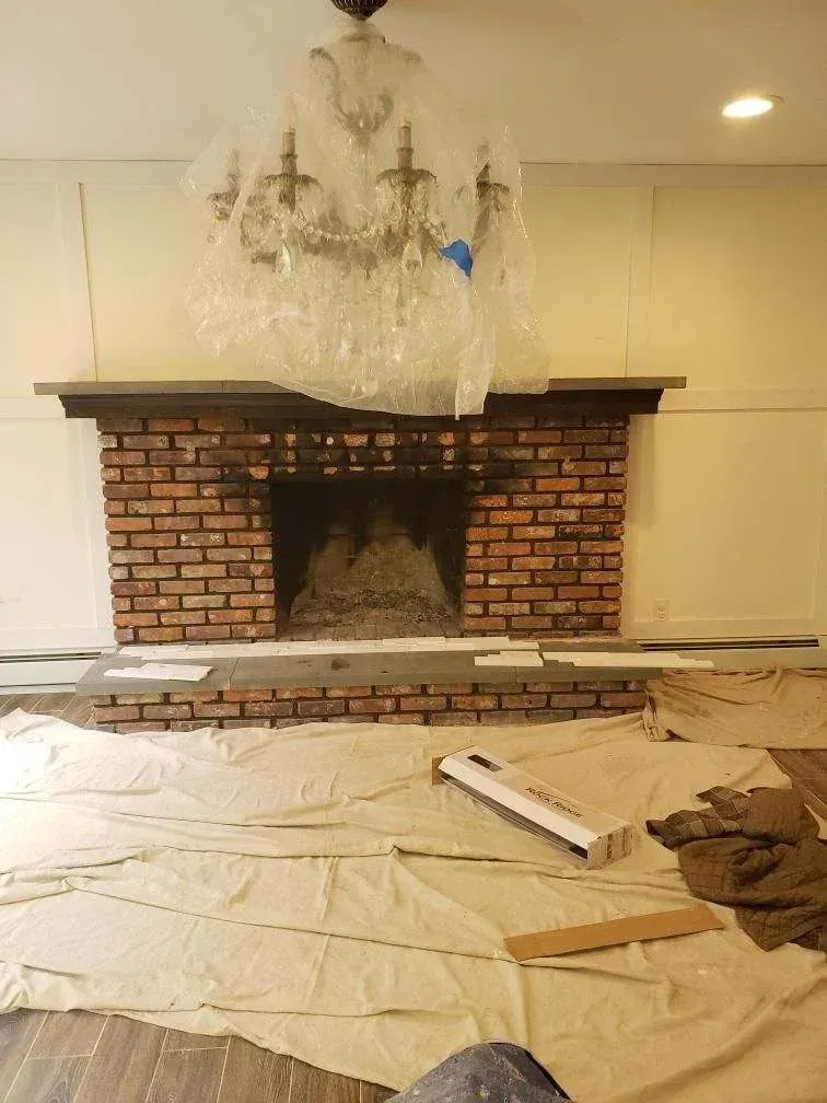 Brick fireplace under renovation, with plastic-covered chandelier and drop cloths on the floor.