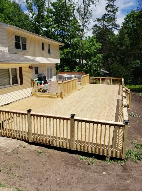 Wooden deck attached to a two-story house; surrounded by railings. Green lawn, trees in the background.