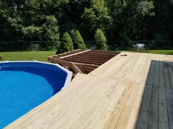 Wooden deck surrounds an above-ground pool. Construction in progress. Trees and green lawn in background.