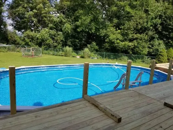 Above-ground pool with wooden deck and posts, set in a grassy backyard with trees.