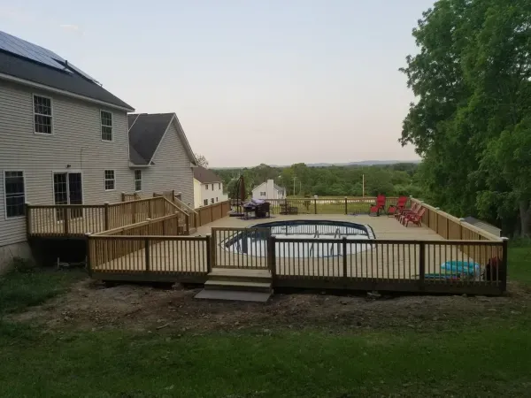 Wooden deck surrounds an above-ground pool, connected to a house with a ramp. Red chairs sit poolside.