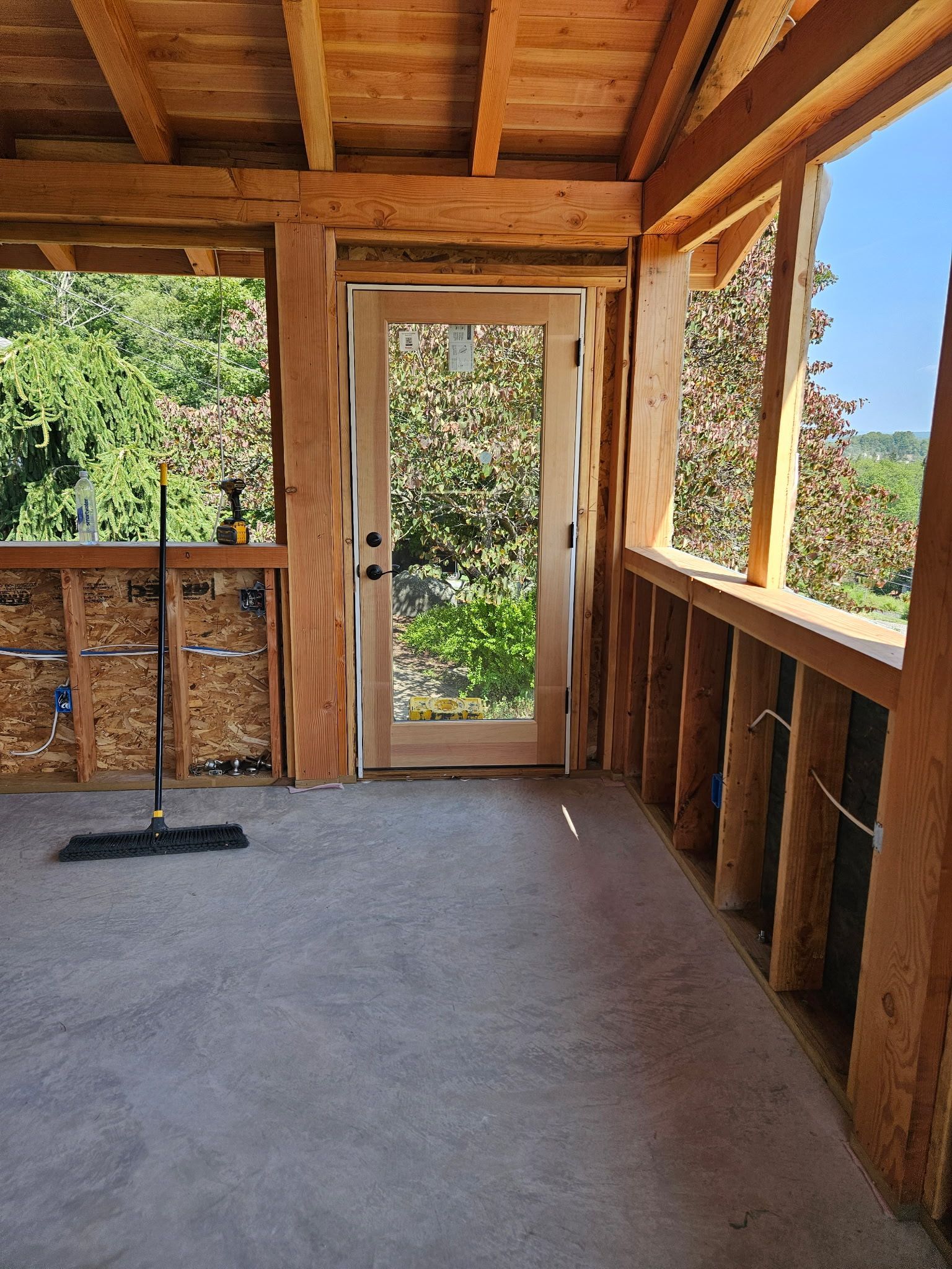Unfinished porch under construction with wooden framing, concrete floor, door, and view of trees.
