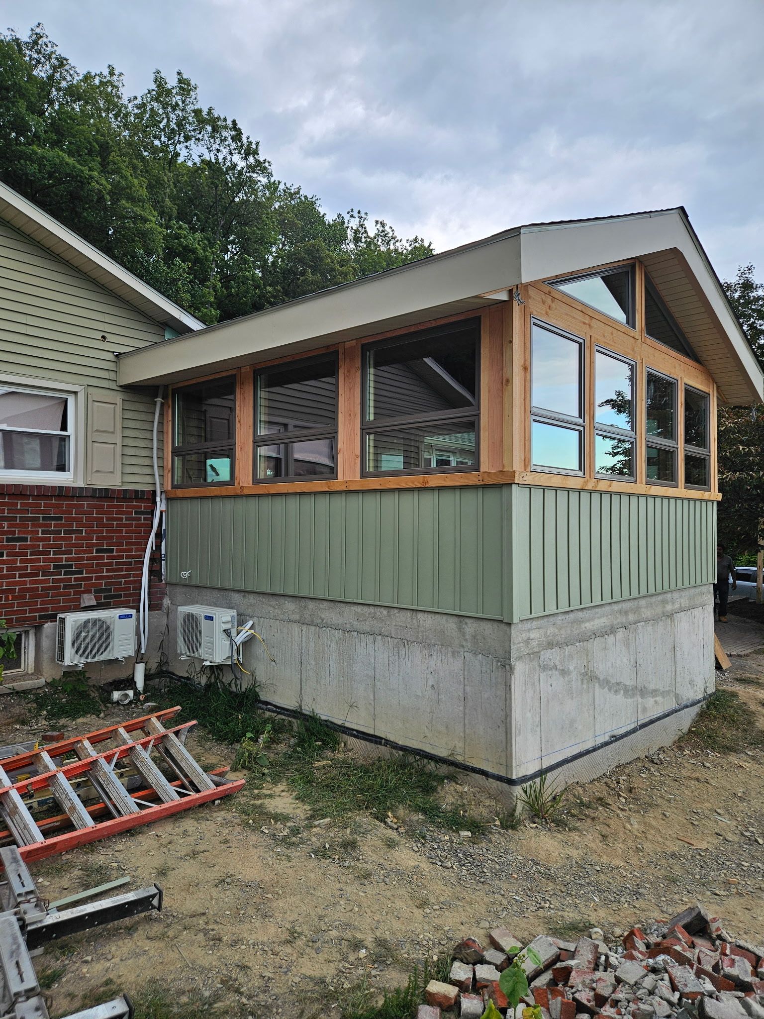 Sunroom addition with light wood trim and large windows, connected to a house with green siding.