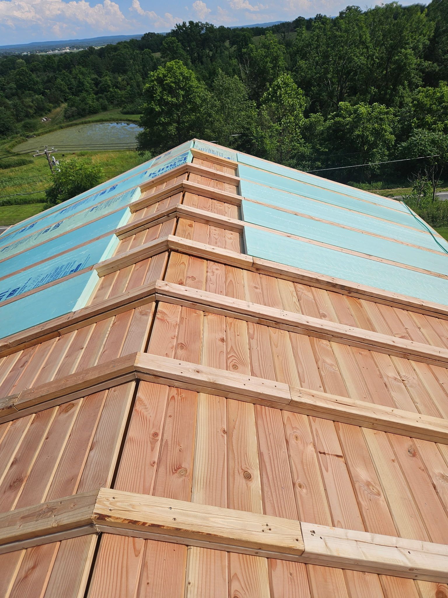 A partially constructed roof with wooden planks and blue insulation, set against a green landscape.