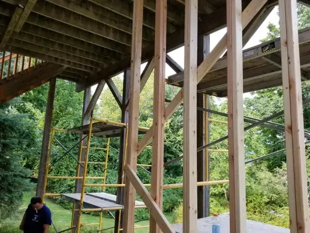 Construction of wooden framing on a deck. Scaffolding and a worker are visible. Green trees in the background.