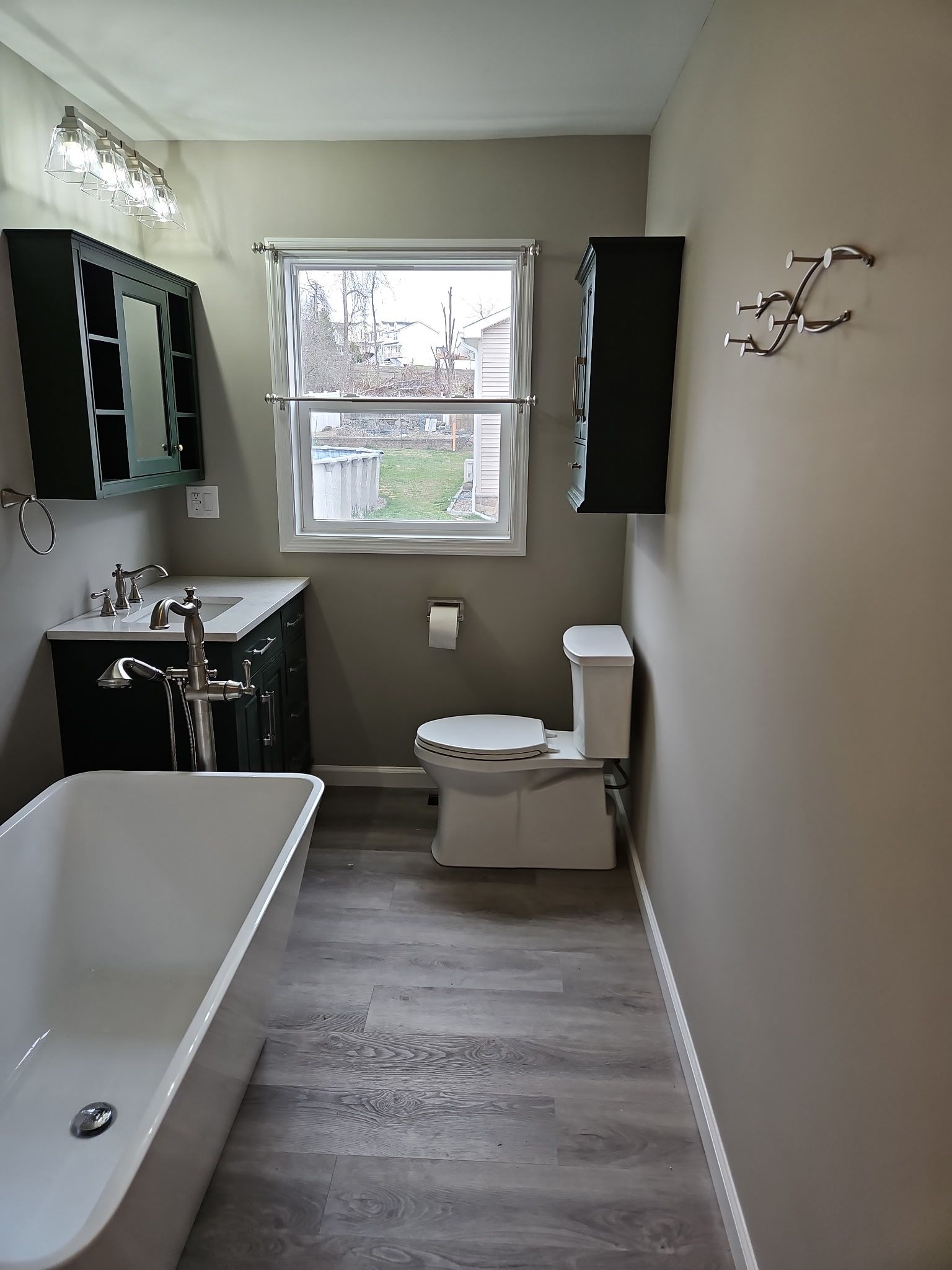 Bathroom with white tub, toilet, dark cabinets, and gray flooring; window with tree view.