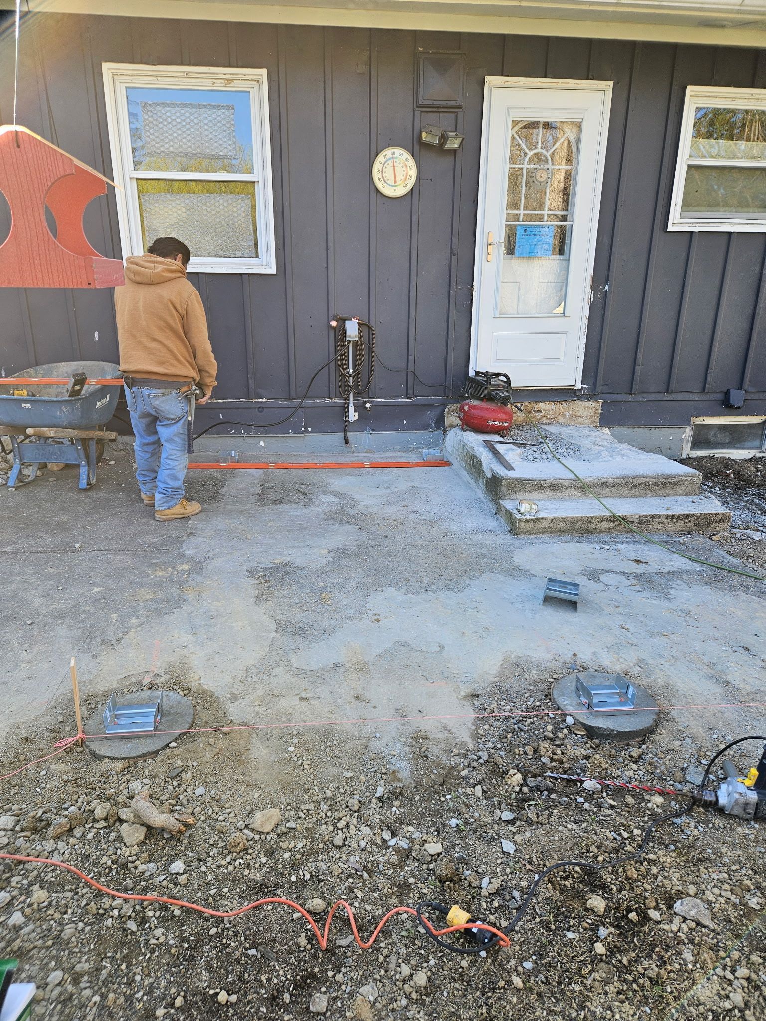 Man working on patio; gray siding, white door and windows, concrete steps, gravel ground, tools visible.