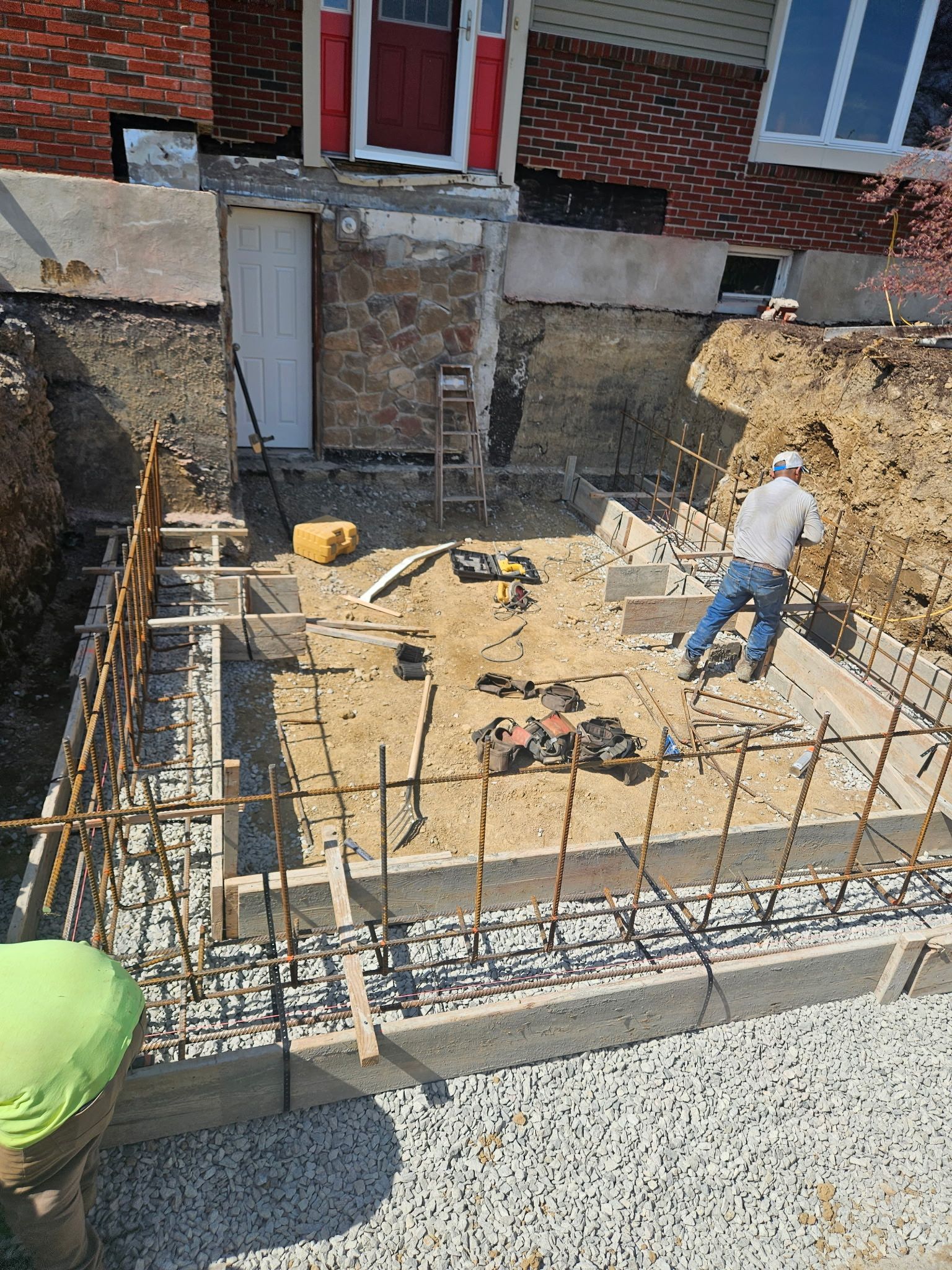 Construction site with workers, rebar, and foundation work near a brick building.