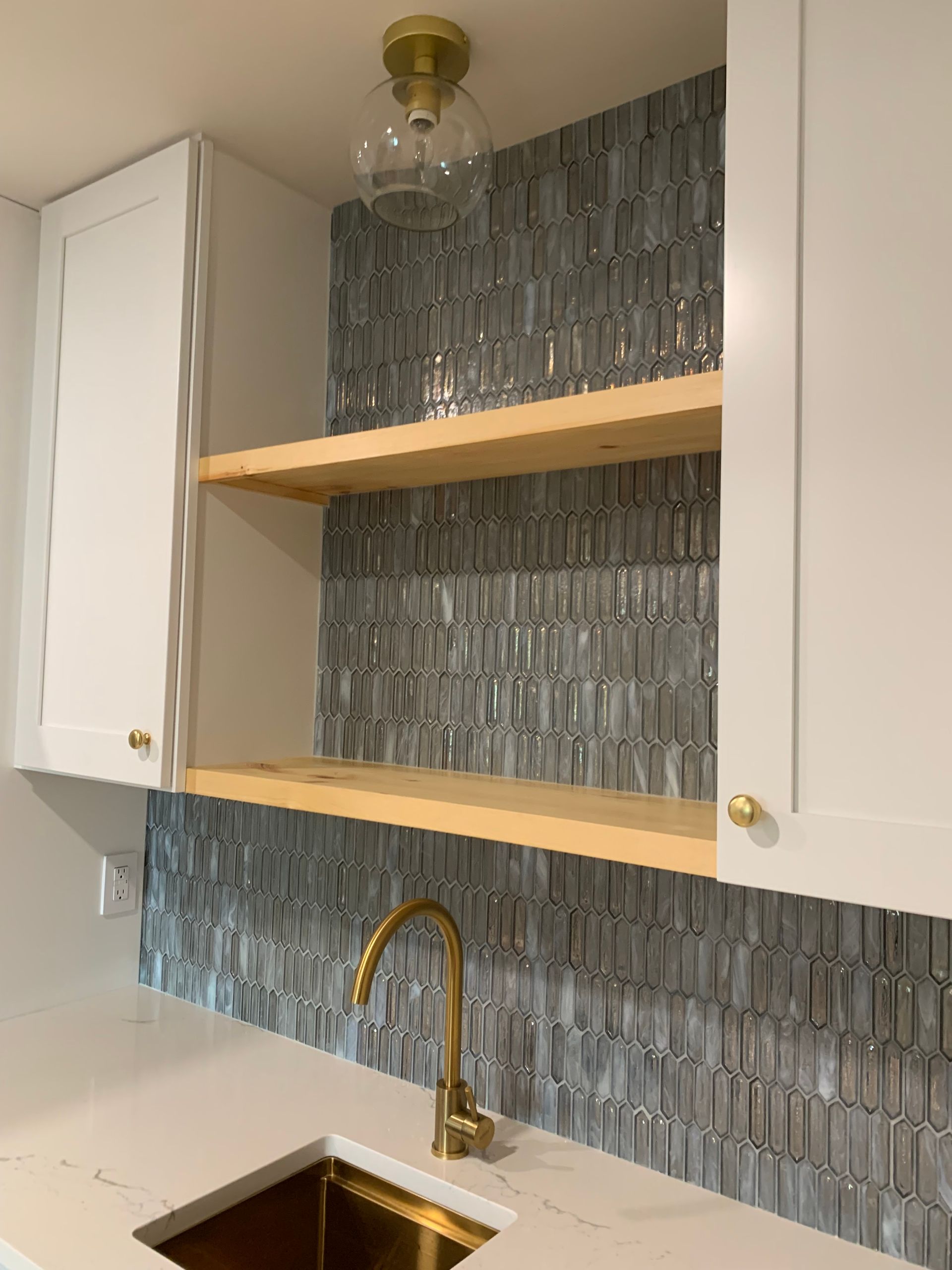 White cabinets flank a bar area with a gold faucet, sink, and backsplash. Wooden shelves are on a tiled wall.