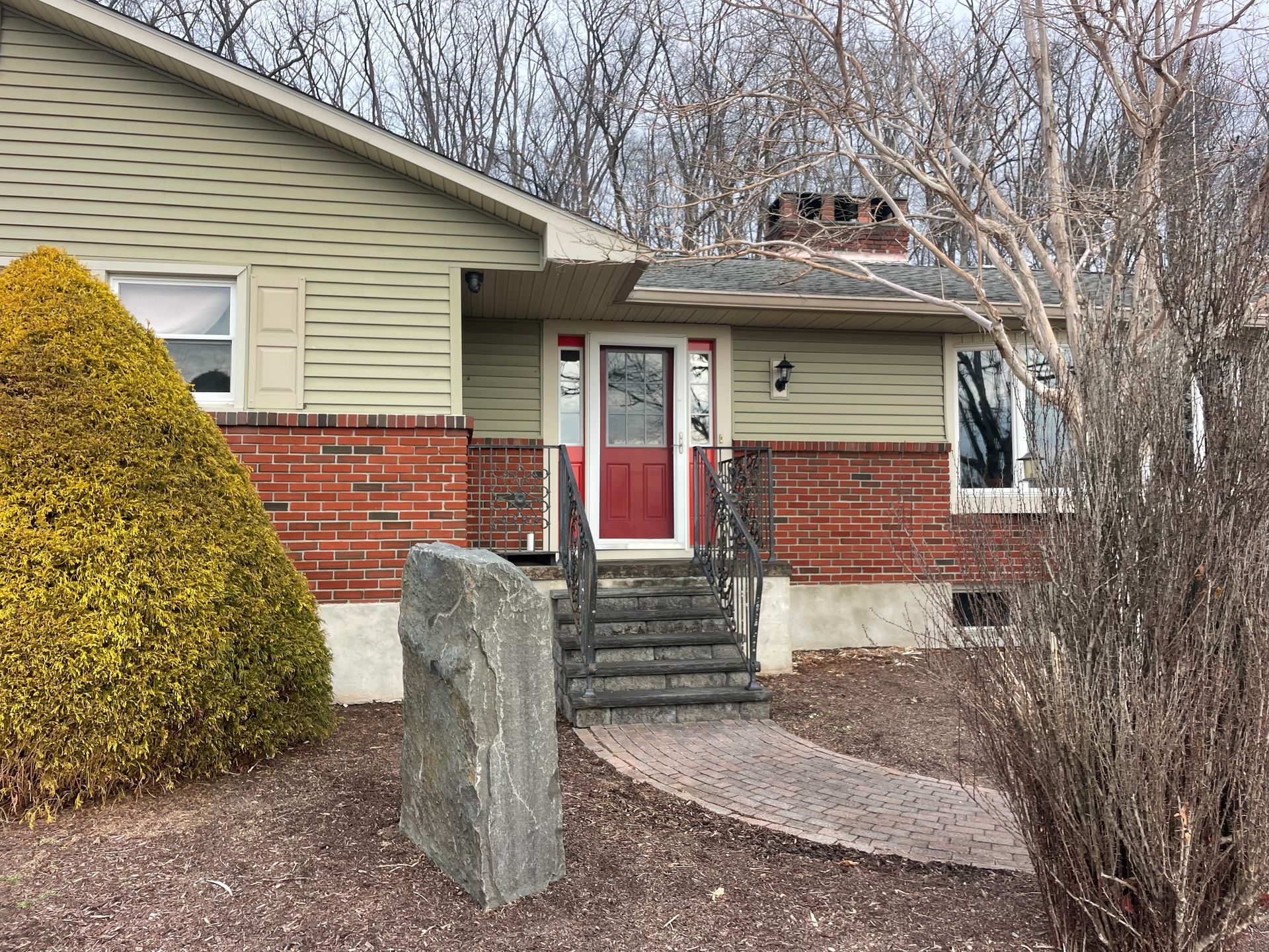 House with red door, brick facade, and stone pathway leading to steps. A large rock stands in the foreground.