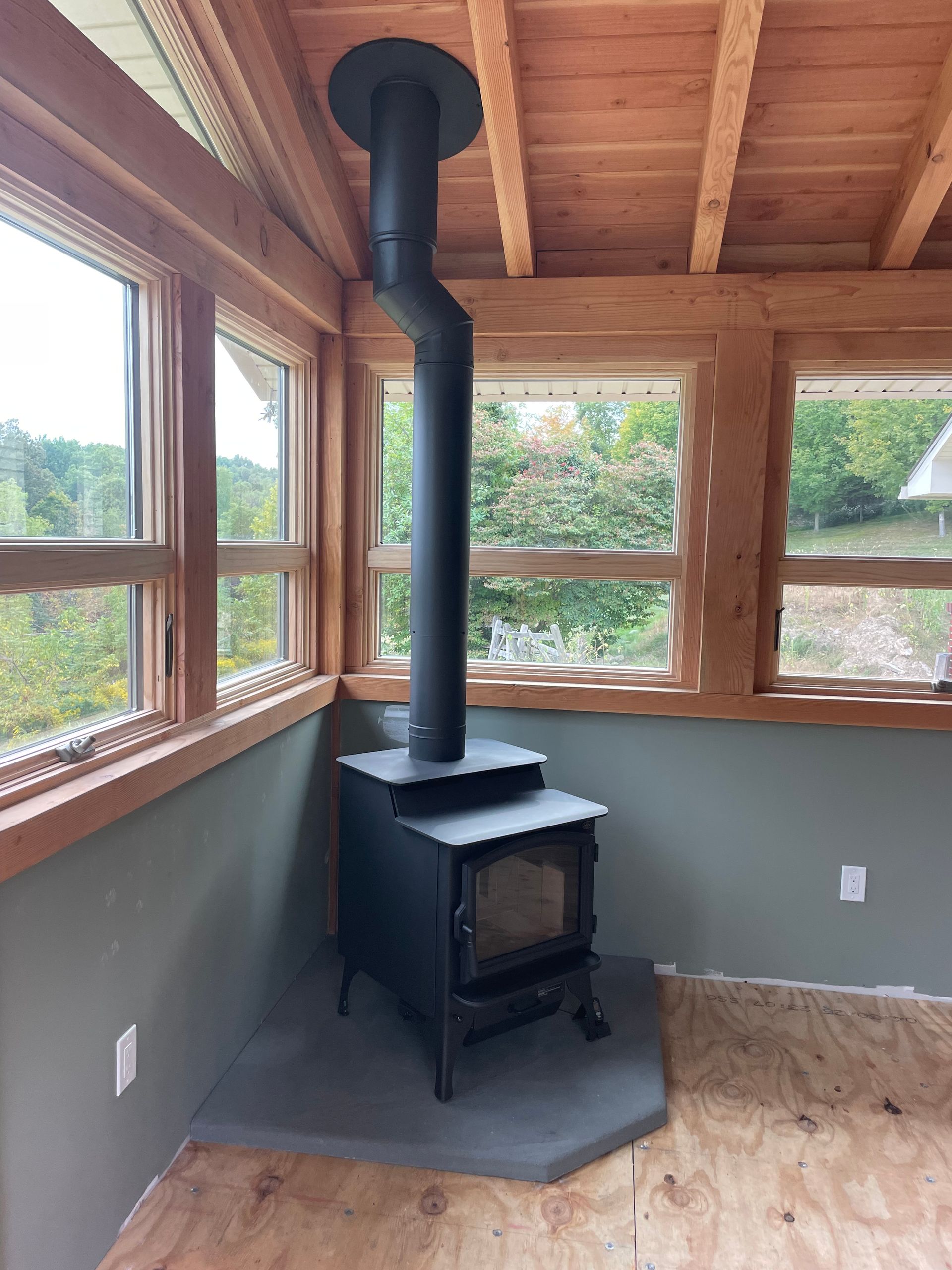 A black wood-burning stove with chimney in a room with wood-framed windows and green walls.