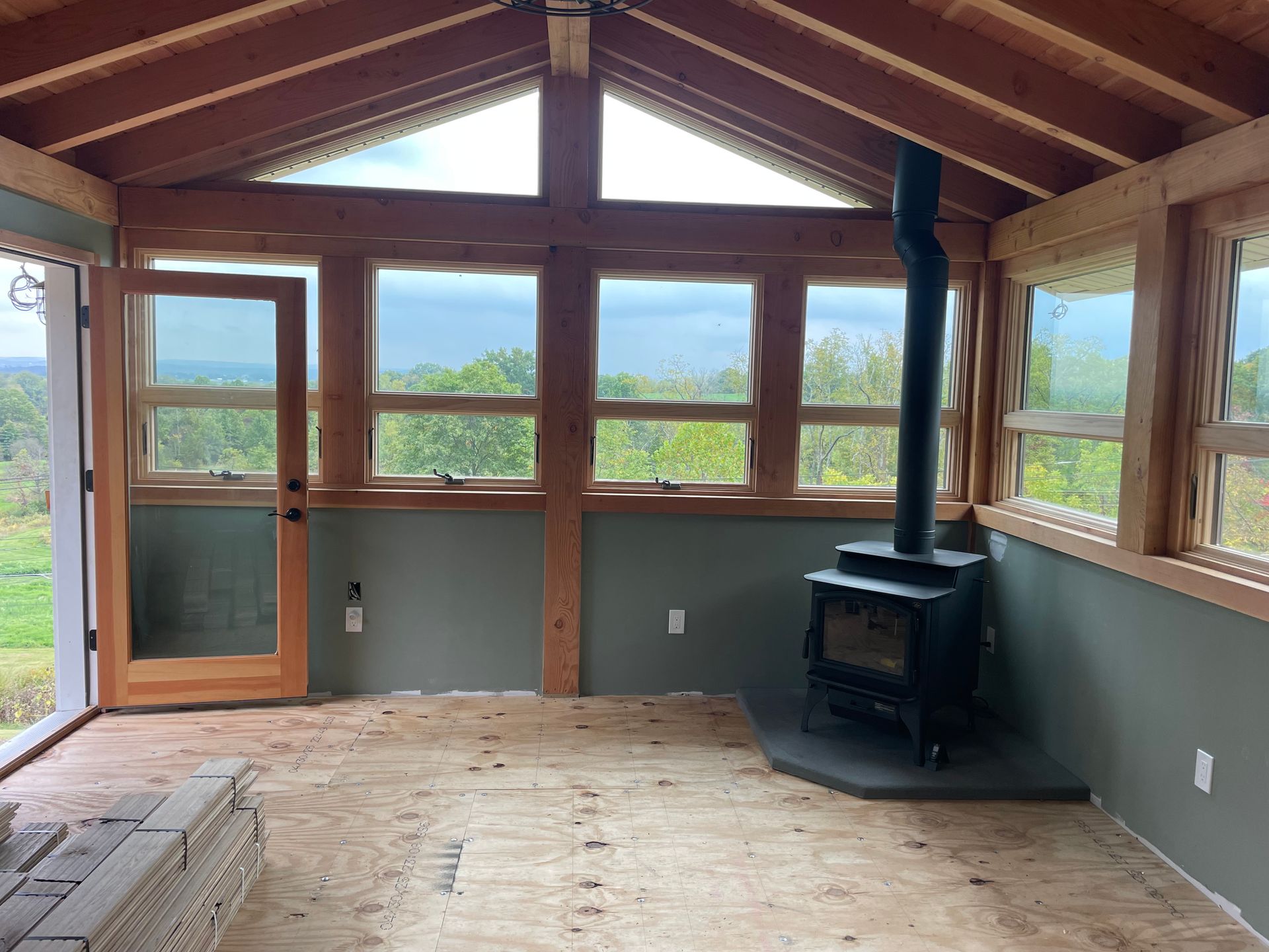 Interior of a sunroom with wood beams, windows, and a wood-burning stove overlooking a green landscape.