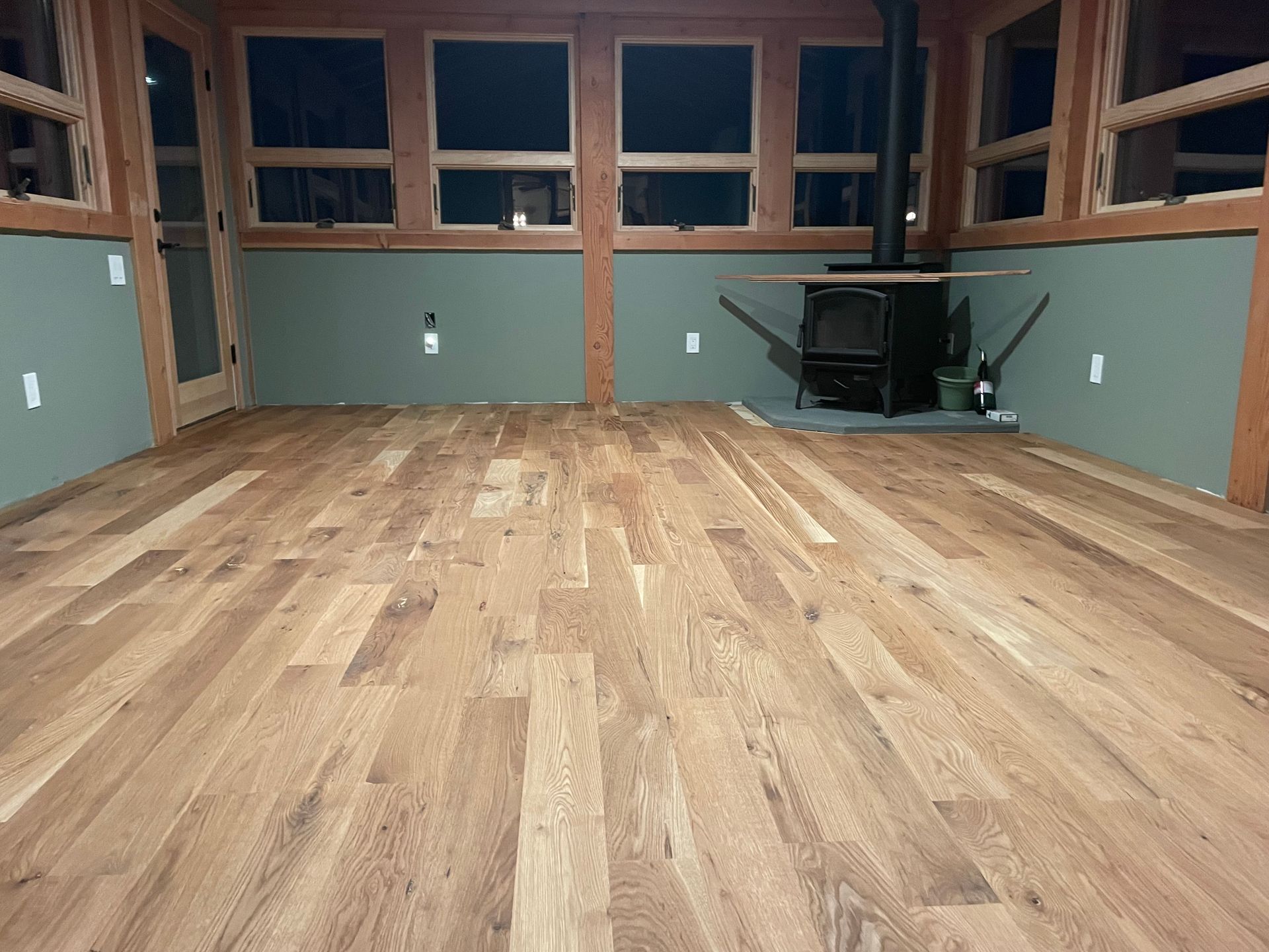 Empty room with wood floor, green walls, wooden window frames, and a wood-burning stove.