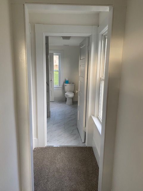 Hallway leading into a bathroom. White walls, doorway, trim. Toilet, window visible at the end.