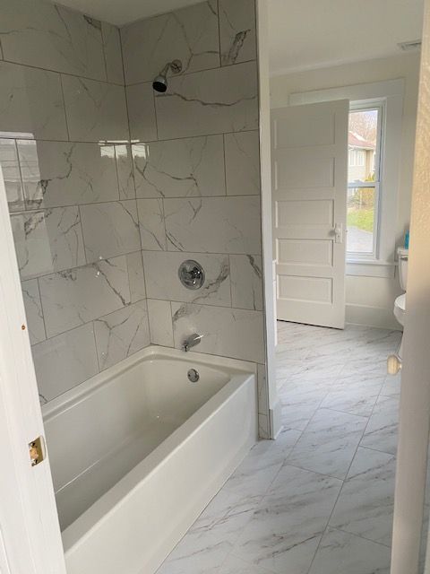 White-tiled bathroom with a bathtub, showerhead, and an open doorway leading to a toilet and window.