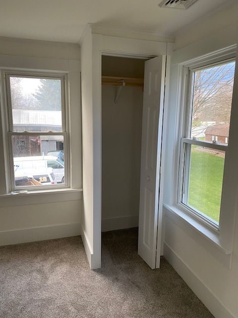 Empty room with closet, windows, and gray carpet. White walls and trim. Sunlight through windows.