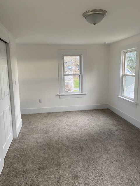 Empty bedroom with gray carpet, white walls, two windows, and a closet door.