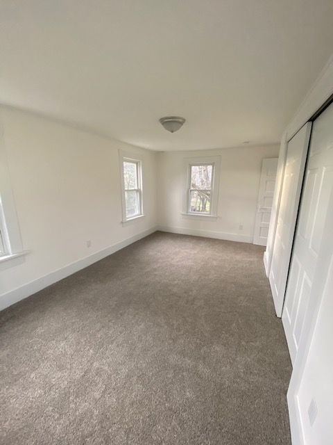 Empty bedroom with tan carpet, white walls, two windows, and closet doors.