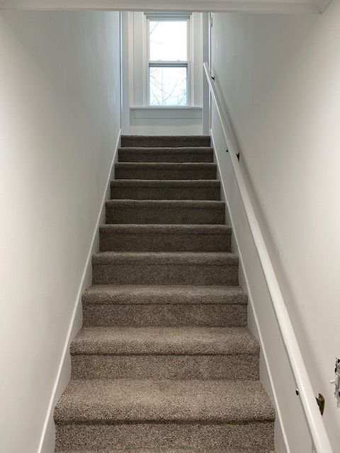 Staircase with beige carpet and white walls, leading up to a window at the top. White handrail on the right.