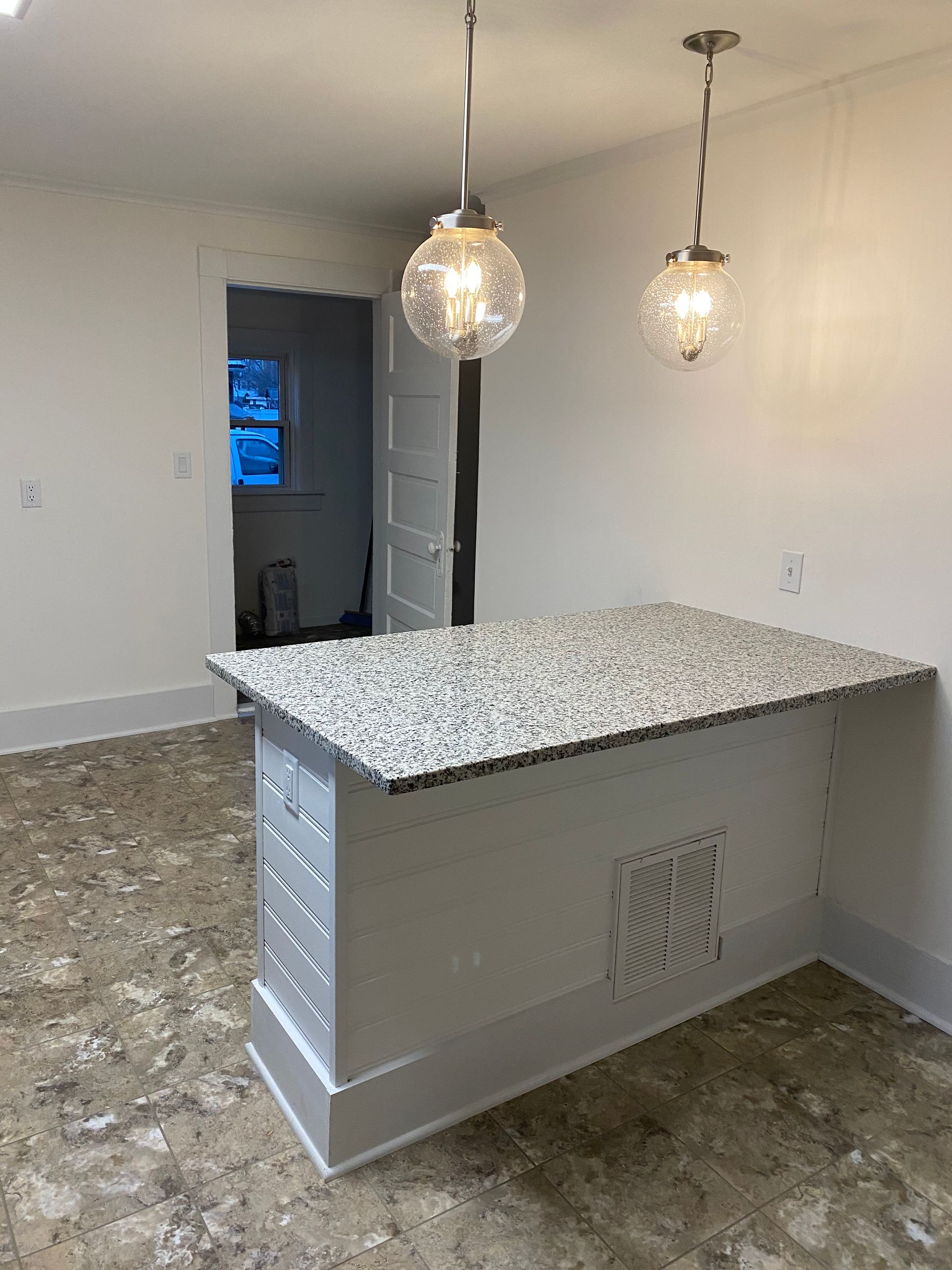 Kitchen island with granite countertop and pendant lights, in a room with white walls and a doorway.