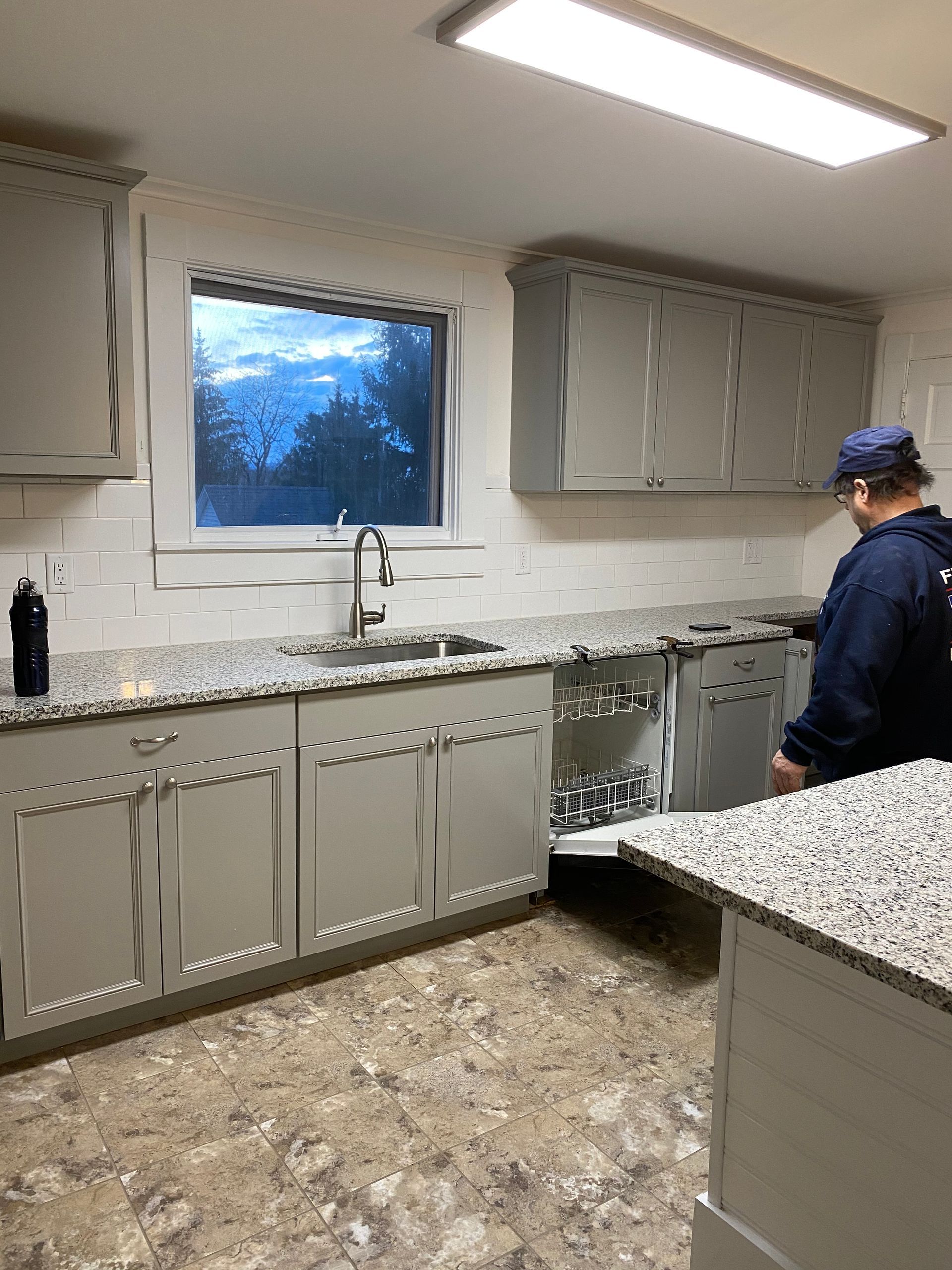 A person installing a dishwasher in a gray and white kitchen with a granite countertop.