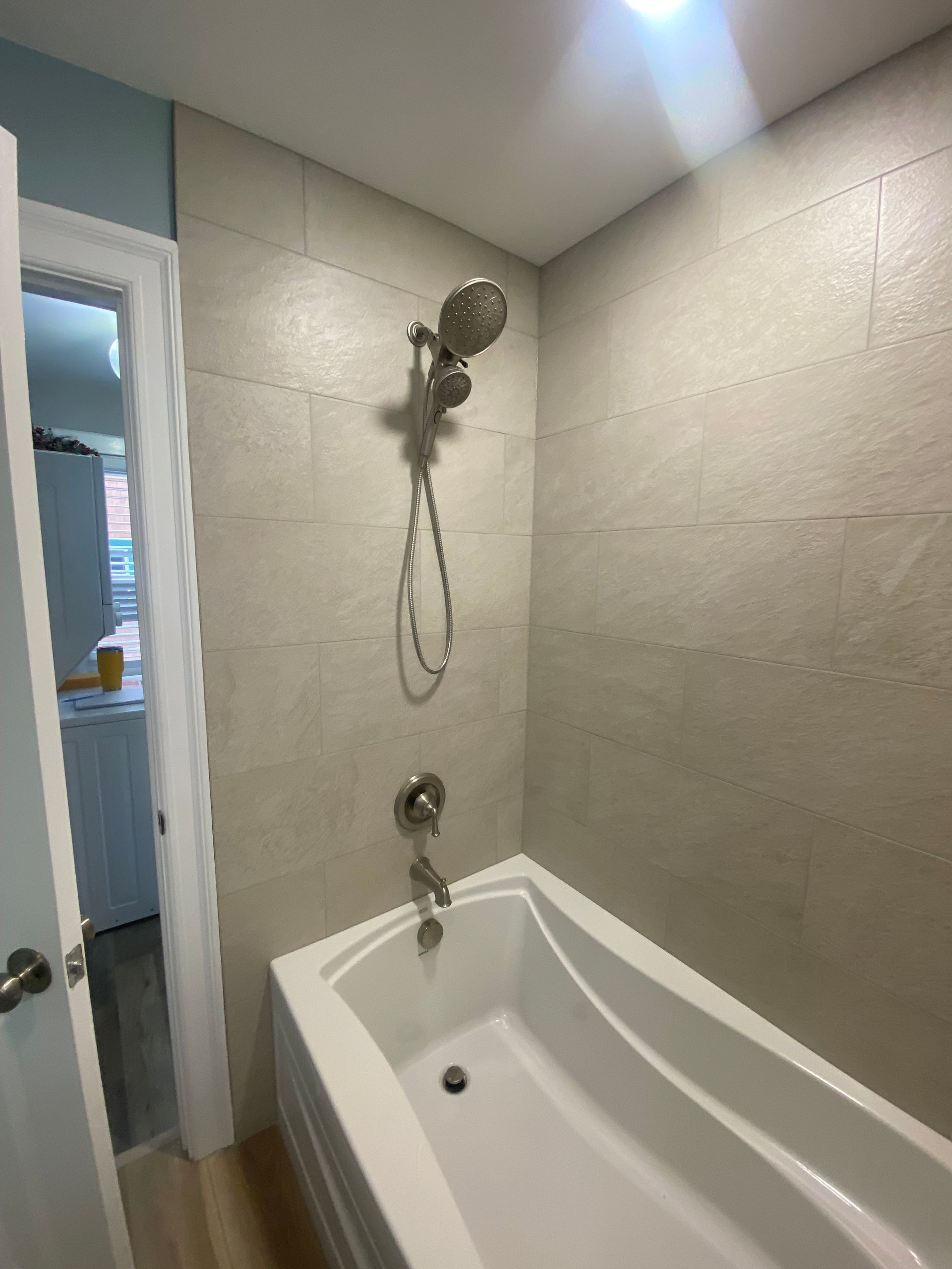 Small bathroom with a white bathtub, tiled walls, and silver shower fixtures. A door is open on the left.