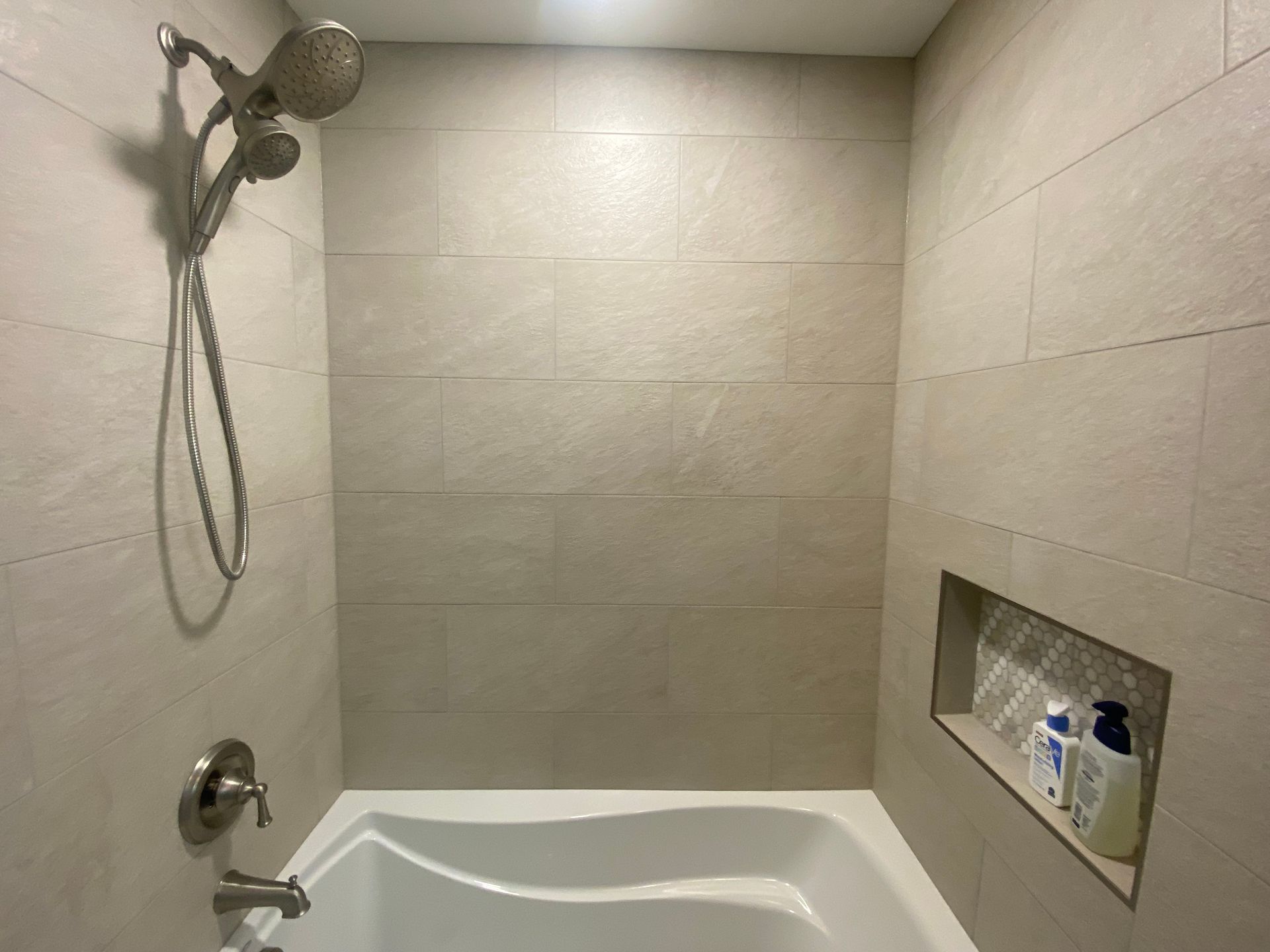 Shower with beige tile walls, white tub, and built-in shelf with toiletries.