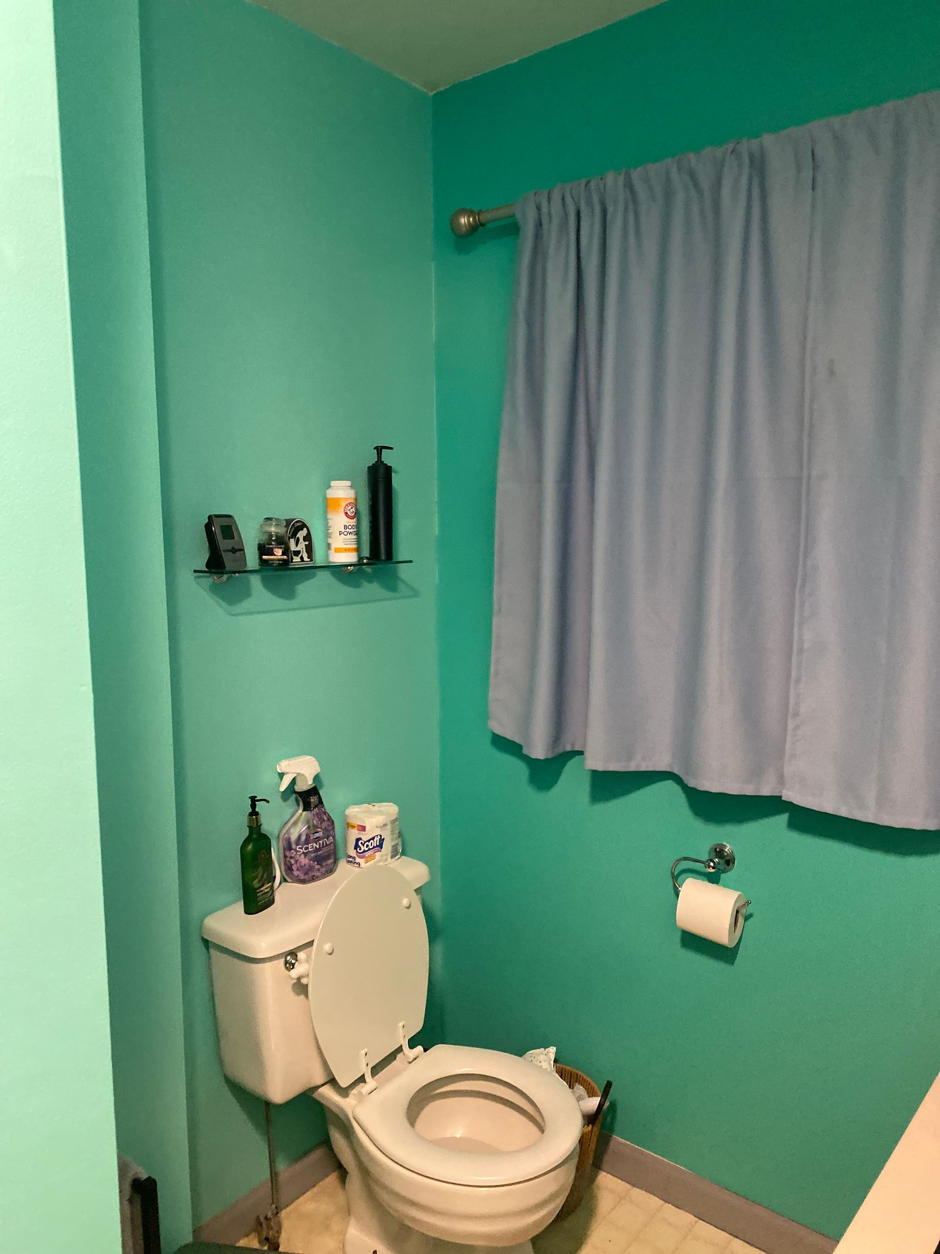 Teal bathroom with white toilet, blue curtain, and various toiletries on a shelf.