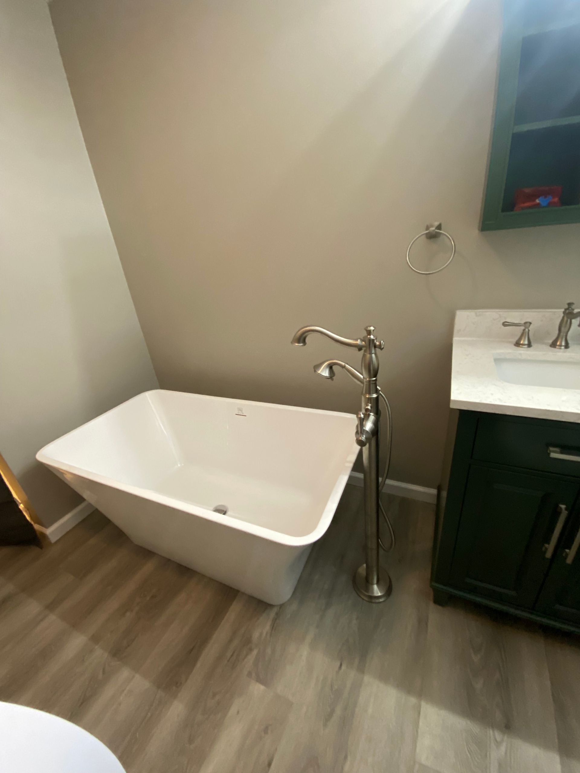 Freestanding white bathtub in a bathroom with green cabinets and light wood-look flooring.