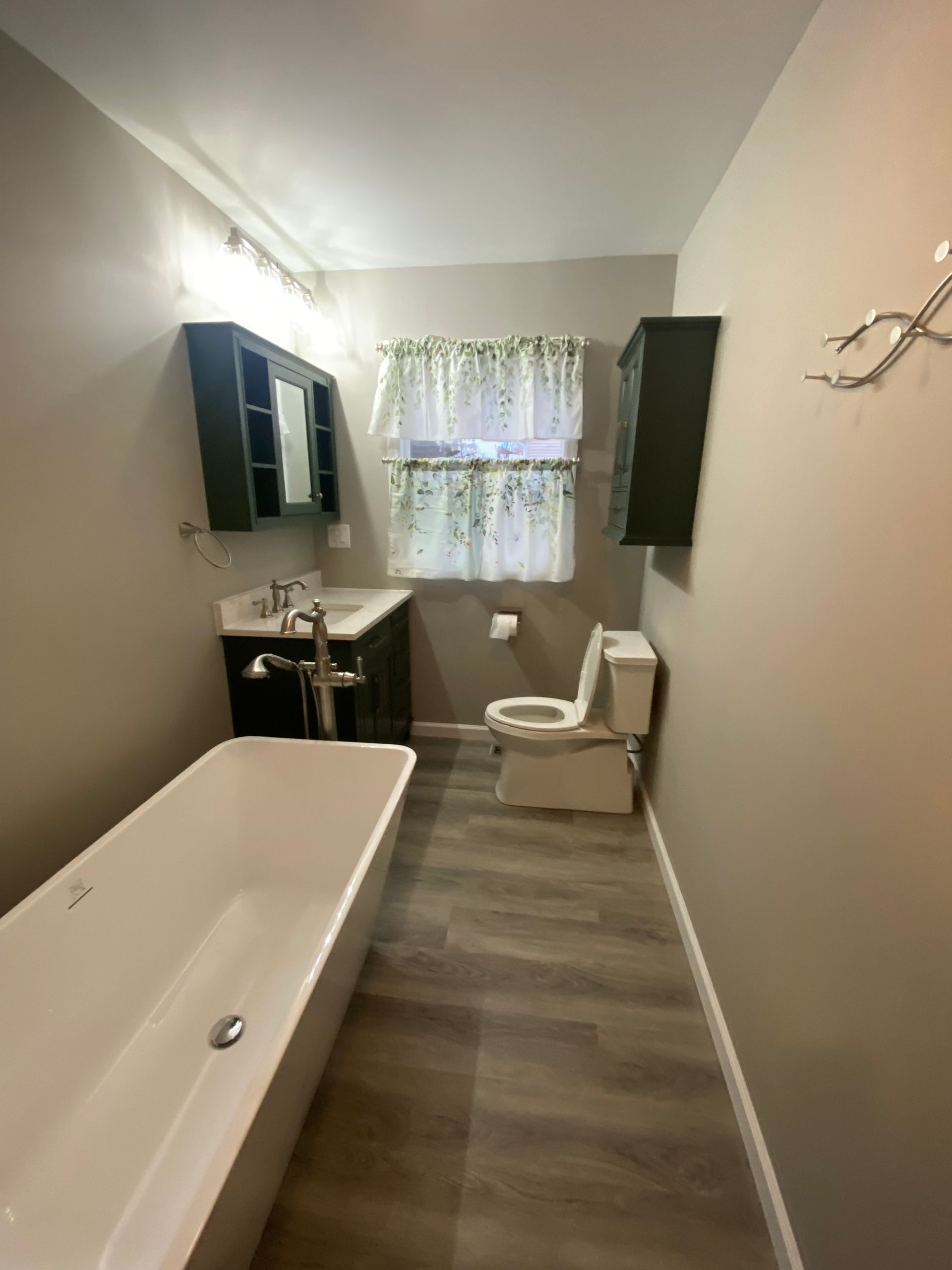 Bathroom with a white tub, dark green vanity, and matching storage. Light green curtain and gray walls.