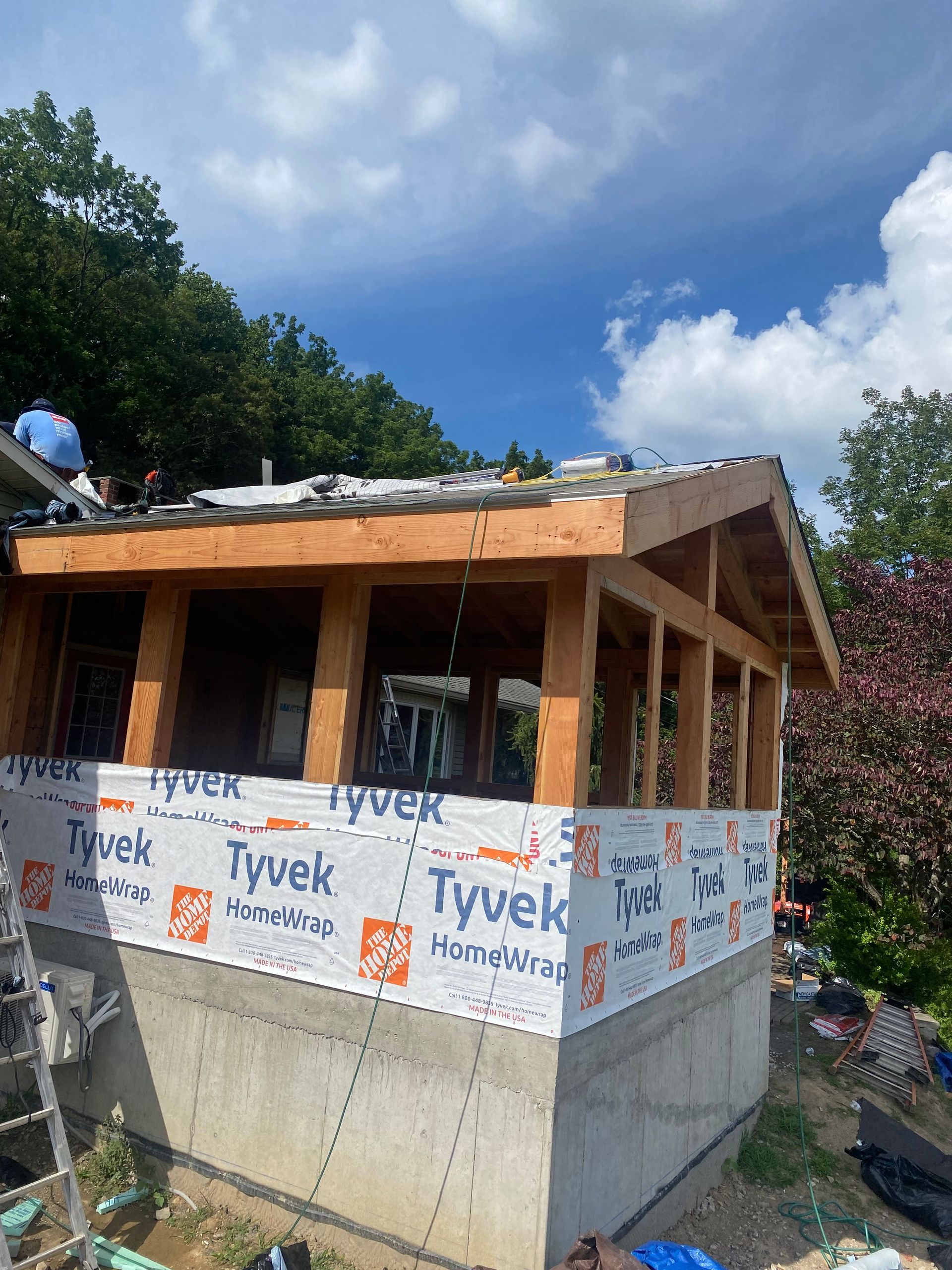 Construction of a small wood-framed building with Tyvek wrap on a concrete foundation, under a partly cloudy sky.