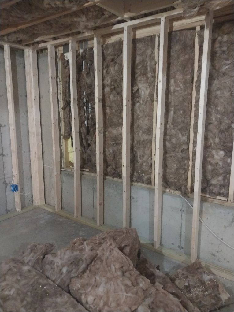 Interior view of a framed wall with insulation, studs, and concrete floor. Construction in progress.