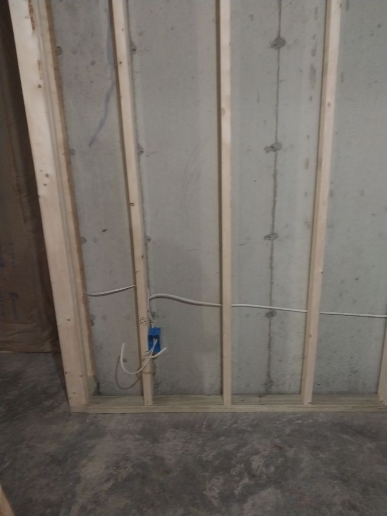 Wooden wall framing with a blue electrical box and wiring against a concrete wall.