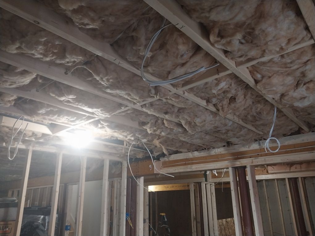 Interior view of a construction site; exposed ceiling beams, insulation, and electrical wires. Wood framing and wall visible.