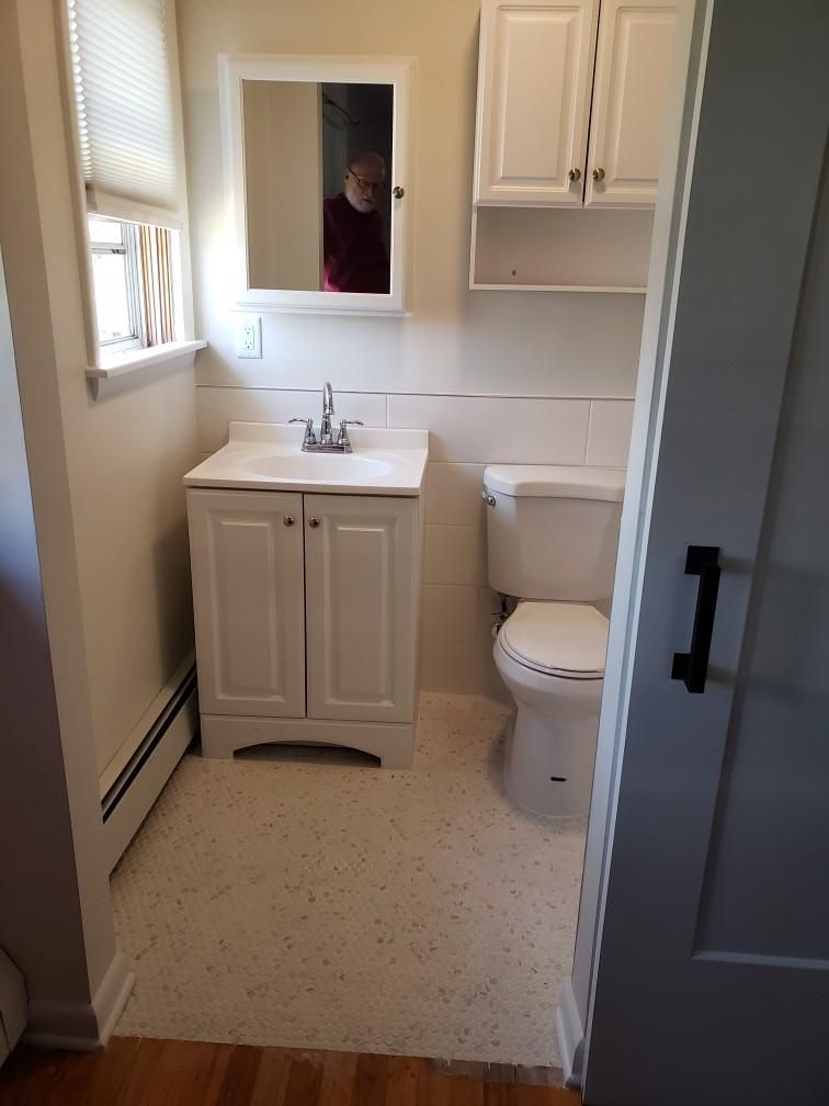 Small white bathroom with vanity, toilet, mirror, and upper cabinet. A person is reflected in the mirror.
