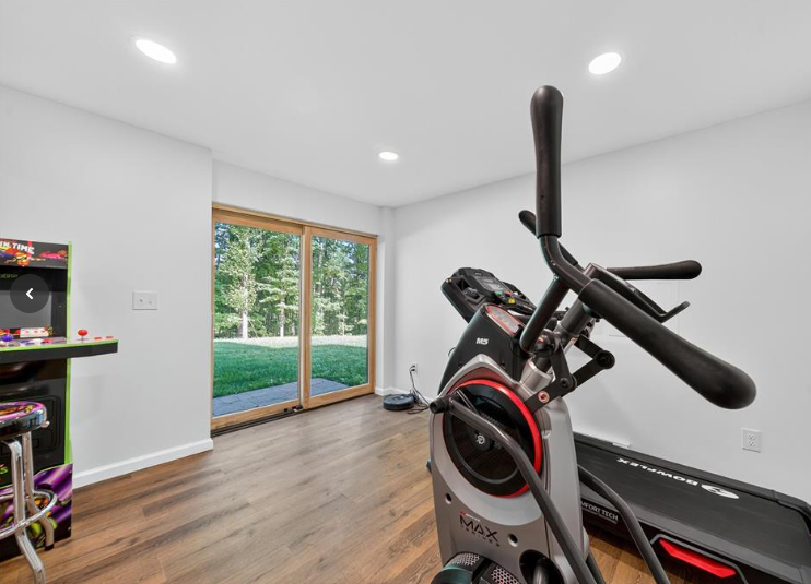 Home gym with elliptical machine, arcade game, and sliding glass doors overlooking a grassy yard.
