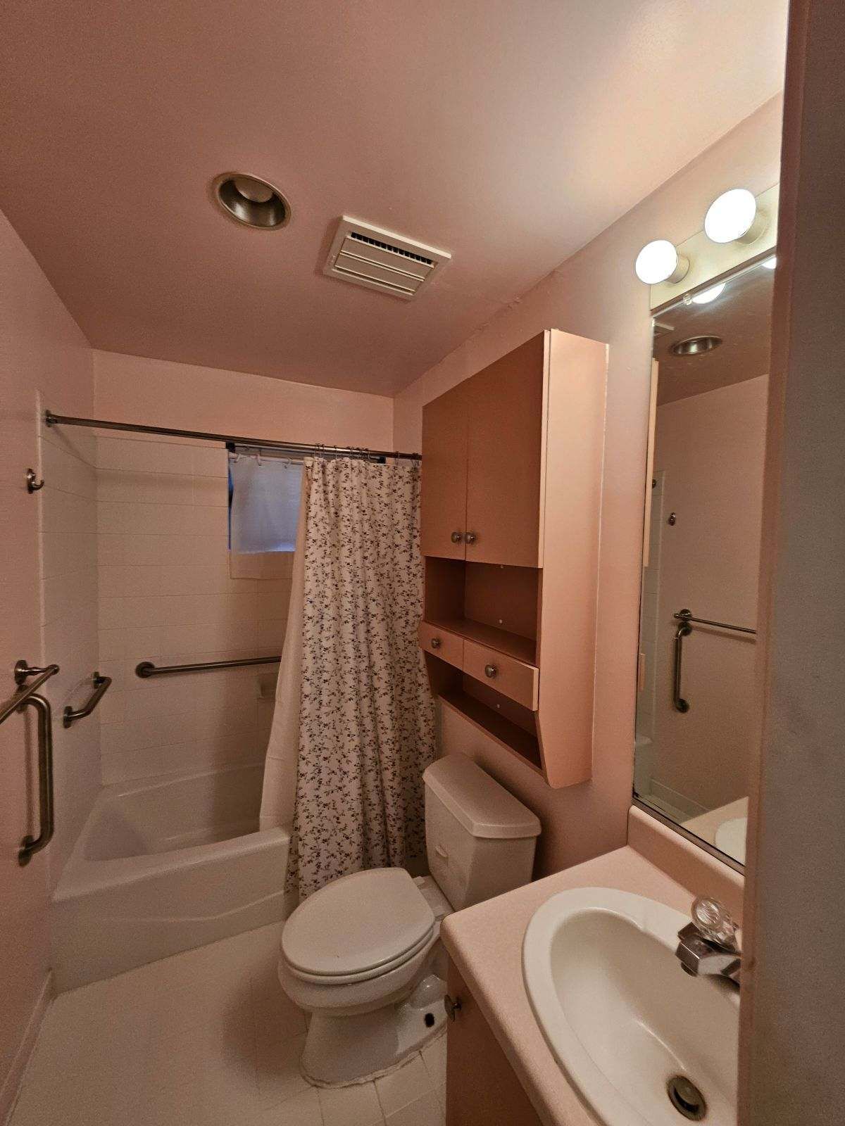 Pink bathroom with a toilet, sink, bathtub, and wall-mounted cabinet. Grab bars and shower curtain visible.