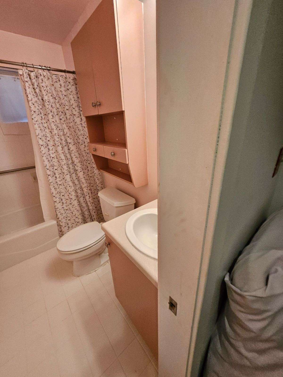 Small bathroom with toilet, sink, and shower/tub. Light pink cabinets and walls; white fixtures.