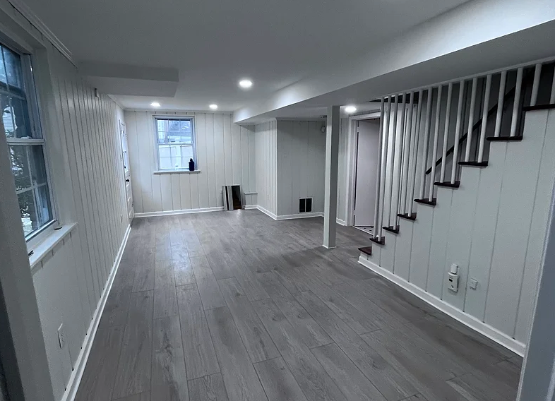 Empty finished basement with gray floors, white paneling, and stairs with a black handrail.