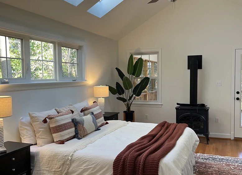 Bedroom with white walls, bed, plant, black stove, windows, and skylights.