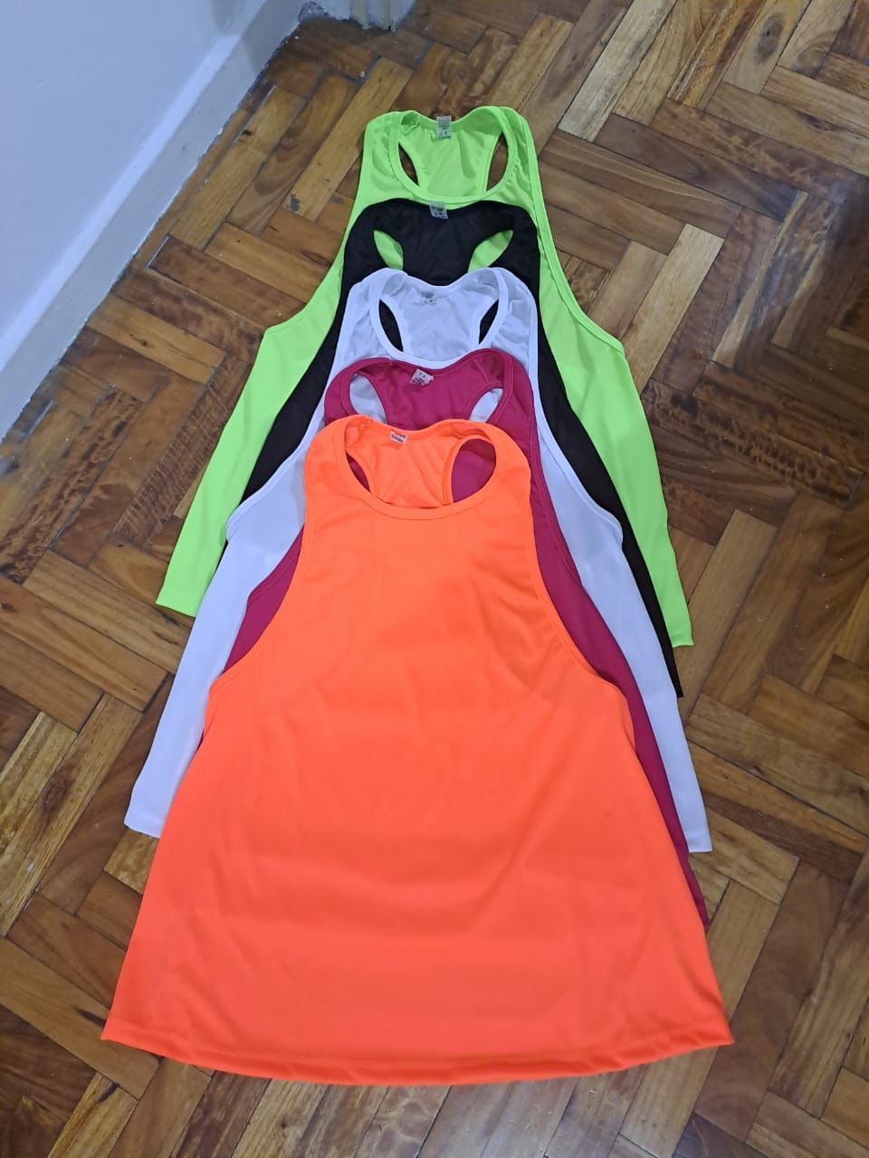 Un montón de camisetas sin mangas de colores neón están apiladas una encima de otra sobre un piso de madera.